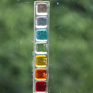 May include: A glass suncatcher with a series of colored squares. The squares are in the colors of the rainbow, starting with red at the bottom and ending with purple at the top.