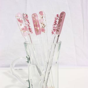 May include: A clear glass mug holds several clear nail art tools. Each tool has a different pink design, including hearts, glitter, and solid pink. The tools are arranged in a fan shape, with the handles visible.