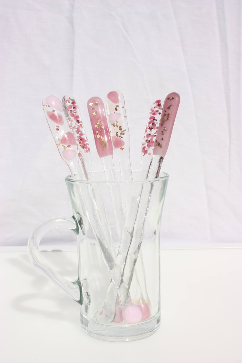 May include: A clear glass mug holds several clear nail art tools. Each tool has a different pink design, including hearts, glitter, and solid pink. The tools are arranged in a fan shape, with the handles visible.