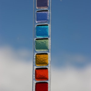 May include: A stained glass suncatcher with a rainbow of colors. The suncatcher is made of glass squares in shades of purple, blue, green, yellow, orange, and red.