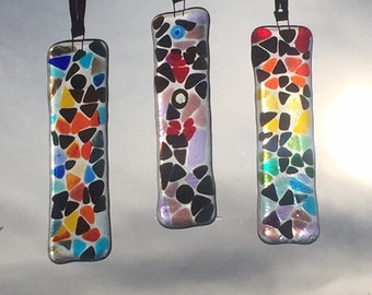 May include: Three colorful glass mosaic suncatchers hanging from a string. Each suncatcher is rectangular and features a different color scheme, including black, white, red, orange, yellow, green, blue, and purple.