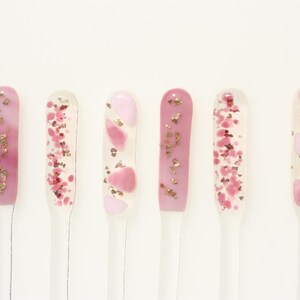 May include: Six nail art samples in a row, each with a different design. The designs feature pink and clear gel with gold and pink glitter and shapes. The nail art samples are on clear sticks against a white background.
