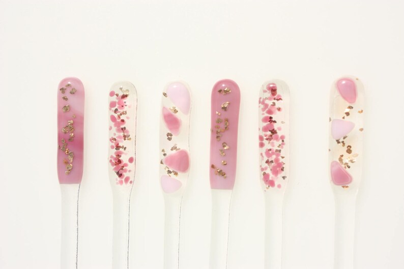 May include: Six nail art samples in a row, each with a different design. The designs feature pink and clear gel with gold and pink glitter and shapes. The nail art samples are on clear sticks against a white background.