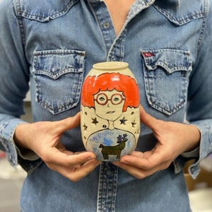 May include: A white ceramic vase with a hand-painted design of a woman with red hair wearing glasses and a blue sweater with a reindeer. The vase has a star-shaped cut-out pattern.
