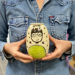 May include: A white ceramic candle holder shaped like a person with a green and yellow striped sweater, black hair, and glasses. The candle holder has a holly sprig on top and stars cut out around the edges.