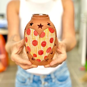 May include: A terracotta vase with a heart-shaped design. The heart is painted with yellow stripes and red polka dots. The vase has star-shaped cutouts around the heart.