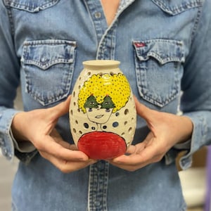 May include: A white ceramic vase with a face painted on it. The face has yellow hair, green eyes, and a red mouth. The vase has holes all over it.