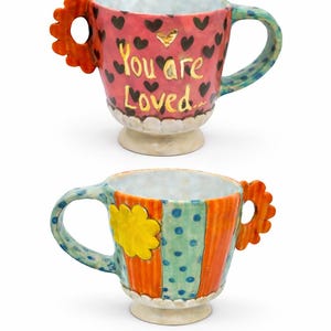 May include: Two brightly coloured ceramic mugs. One is pink with black hearts and the words "You are Loved". The other is orange, blue, and green with a yellow flower. Both have unique handles and decorative bases.
