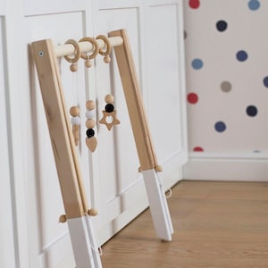 Wooden baby gym made of alder wood, bottoms of the legs are painted white. Legs are connected with white strings and wooden beads. Hanging toys made of wooden and crochet beads in white and black color.  Two heart and star shapes on hangers.