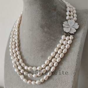 May include: A three-strand pearl necklace with a silver flower clasp. The pearls are white and have a slightly irregular shape.