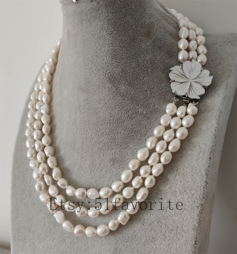 May include: A three-strand pearl necklace with a silver flower clasp. The pearls are white and have a slightly irregular shape.
