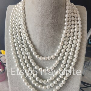 Puede incluir: Un collar de perlas de múltiples hilos con cinco filas de perlas blancas. El collar se muestra en un busto de maniquí blanco.