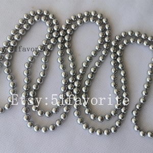 Puede incluir: Varias hebras de collares de cuentas plateadas se disponen sobre una superficie blanca. Las cuentas son pequeñas y redondas, creando una superficie lisa y reflectante. Probablemente sean joyas.