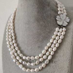 May include: A three-strand pearl necklace with a white flower pendant. The necklace is made of white freshwater pearls and is approximately 18 inches long. The flower pendant is made of mother-of-pearl and has a silver clasp.