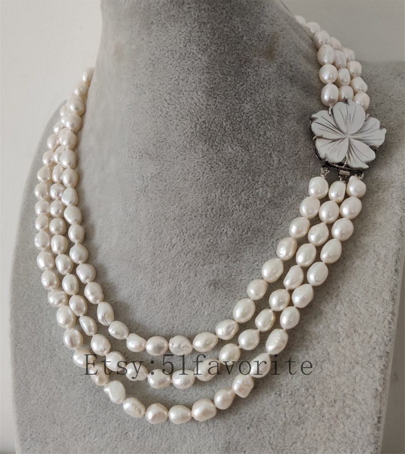 May include: A three-strand pearl necklace with a white flower pendant. The necklace is made of white freshwater pearls and is approximately 18 inches long. The flower pendant is made of mother-of-pearl and has a silver clasp.