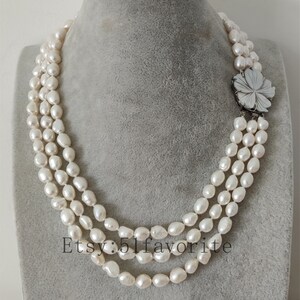 May include: A three-strand pearl necklace with a white flower-shaped clasp. The pearls are white and have a slightly irregular shape.