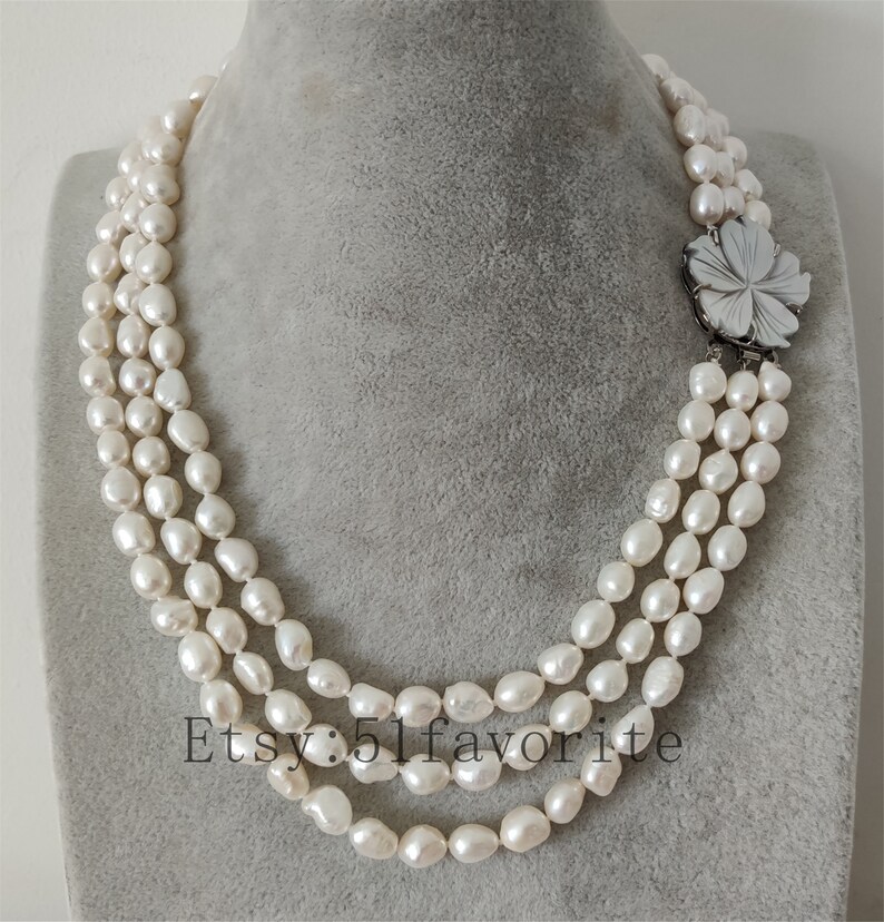 May include: A three-strand pearl necklace with a white flower-shaped clasp. The pearls are white and have a slightly irregular shape.