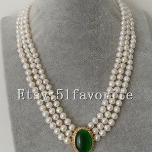 May include: A three-strand pearl necklace with a green gemstone pendant. The pendant is oval-shaped and has a smaller teardrop-shaped green gemstone hanging from it. The necklace is on a white mannequin bust. The earrings are also green gemstones with a gold setting.
