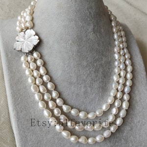 May include: A three-strand pearl necklace with a white flower-shaped clasp. The pearls are white and have a slightly irregular shape.