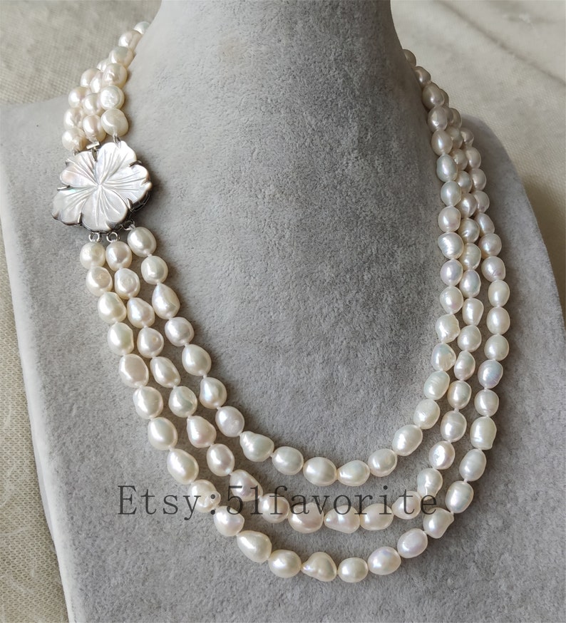 May include: A three-strand pearl necklace with a white flower-shaped clasp. The pearls are white and have a slightly irregular shape.