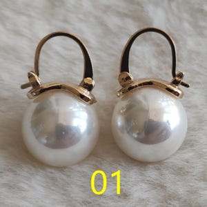May include: A pair of gold-toned hoop earrings featuring large, spherical white faux pearls. The metal hoops attach to the top of each pearl with a curved, polished gold-coloured setting. The number 01 is printed in yellow at the bottom centre.
