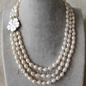 May include: A three-strand pearl necklace with a silver flower clasp. The pearls are white and have a slightly irregular shape.