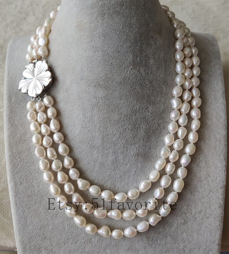 May include: A three-strand pearl necklace with a silver flower clasp. The pearls are white and have a slightly irregular shape.