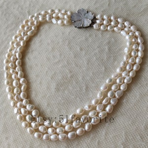 May include: A three-strand pearl necklace with a carved flower clasp. The pearls are white and have a slightly irregular shape. The necklace is approximately 18 inches long.