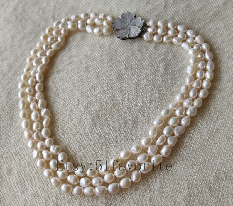 May include: A three-strand pearl necklace with a carved flower clasp. The pearls are white and have a slightly irregular shape. The necklace is approximately 18 inches long.