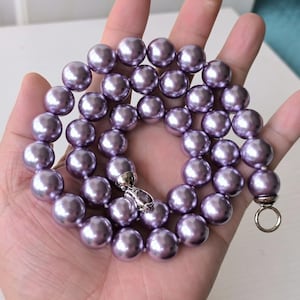 May include: A lavender pearl necklace with a silver clasp and ring closure. The large, round pearls have a lustrous finish. The necklace is displayed on a hand.