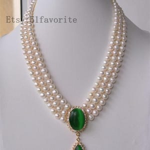 May include: A three-strand pearl necklace featuring a large oval green gemstone pendant. The pendant is encircled by a halo of small clear stones and a smaller teardrop-shaped green gemstone. The necklace is displayed on a white mannequin.