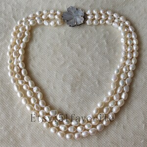 May include: A three-strand pearl necklace with a white flower-shaped clasp. The pearls are white and have a slightly irregular shape.