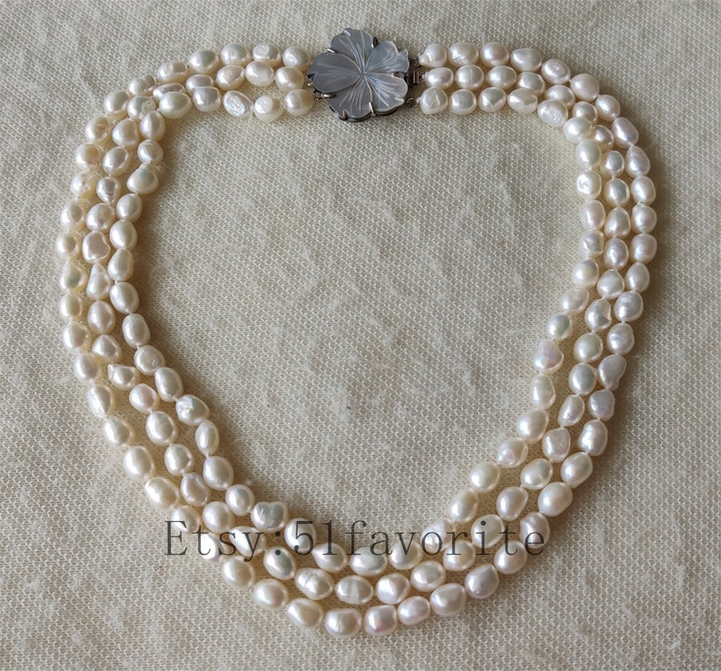 May include: A three-strand pearl necklace with a white flower-shaped clasp. The pearls are white and have a slightly irregular shape.
