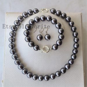 Op de afbeelding: Een sieradenset bestaande uit een ketting, een armband en een paar hangende oorbellen, allemaal voorzien van ronde, donkergrijze kralen met een metallic afwerking. De ketting en armband hebben zilverkleurige, ronde veerringsluitingen.