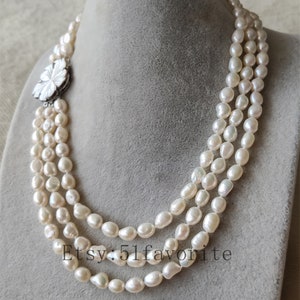 May include: A three-strand pearl necklace with a white flower-shaped clasp. The pearls are white and have a slightly irregular shape.