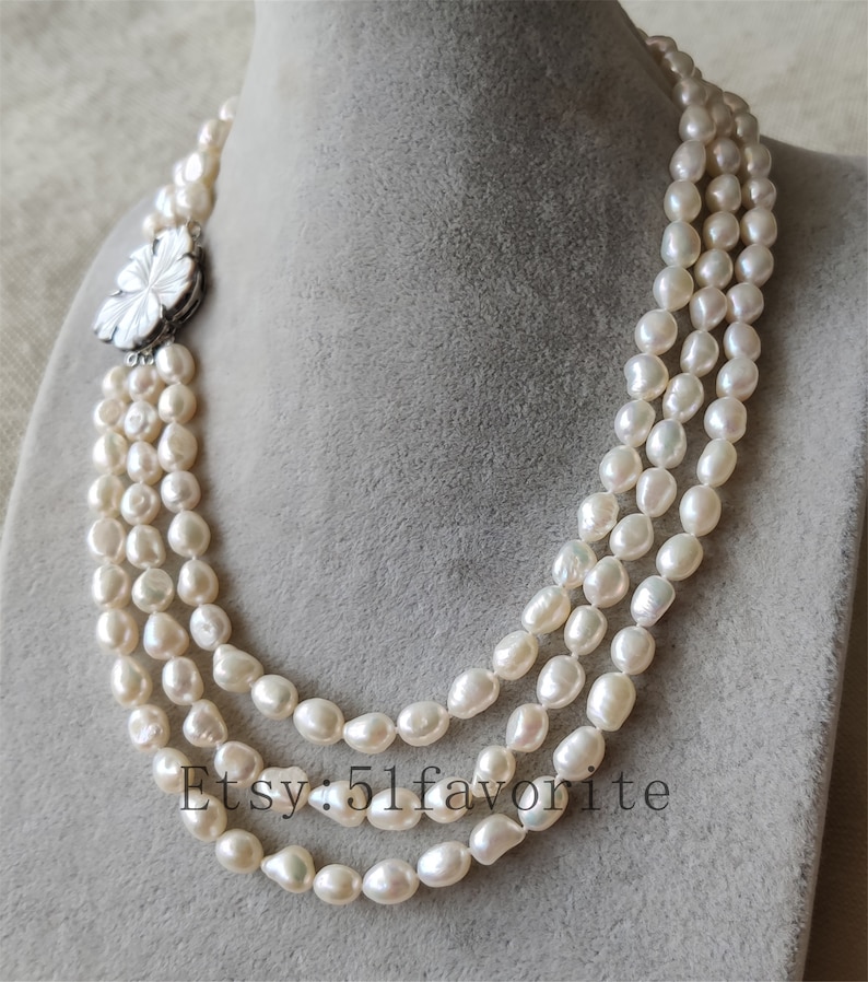 May include: A three-strand pearl necklace with a white flower-shaped clasp. The pearls are white and have a slightly irregular shape.