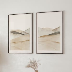 Puede incluir: Dos pinturas abstractas de paisajes enmarcadas en un estilo acuarela, en tonos de marrón, beige y gris. Las pinturas están colgadas en una pared blanca sobre una mesa de madera con sillas.
