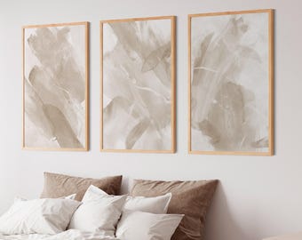 May include: Three framed abstract art prints in neutral tones hang above a bed with white and brown bedding. The artwork features brushstroke-style designs in shades of beige and grey. The room has a minimalist, modern aesthetic.