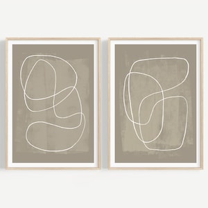 May include: Two framed abstract art prints. Each print features a white continuous line drawing on a muted taupe background. The frames are a light wood color, and the overall aesthetic is minimalist and modern.