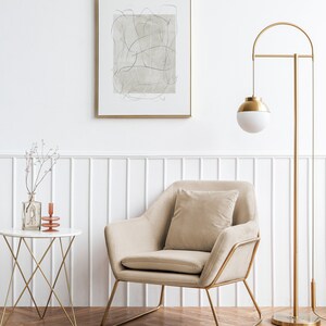 K&ouml;nnte beinhalten: Ein beiger Sessel mit goldenem Metallrahmen und einem beigen Kissen steht in einem Raum mit wei&szlig;en W&auml;nden und einer goldenen Stehlampe mit wei&szlig;em Globus. Ein kleiner wei&szlig;er Tisch mit goldenem Metallrahmen steht im Vordergrund mit einer Vase mit getrockneten Zweigen und einem Kerzenhalter.