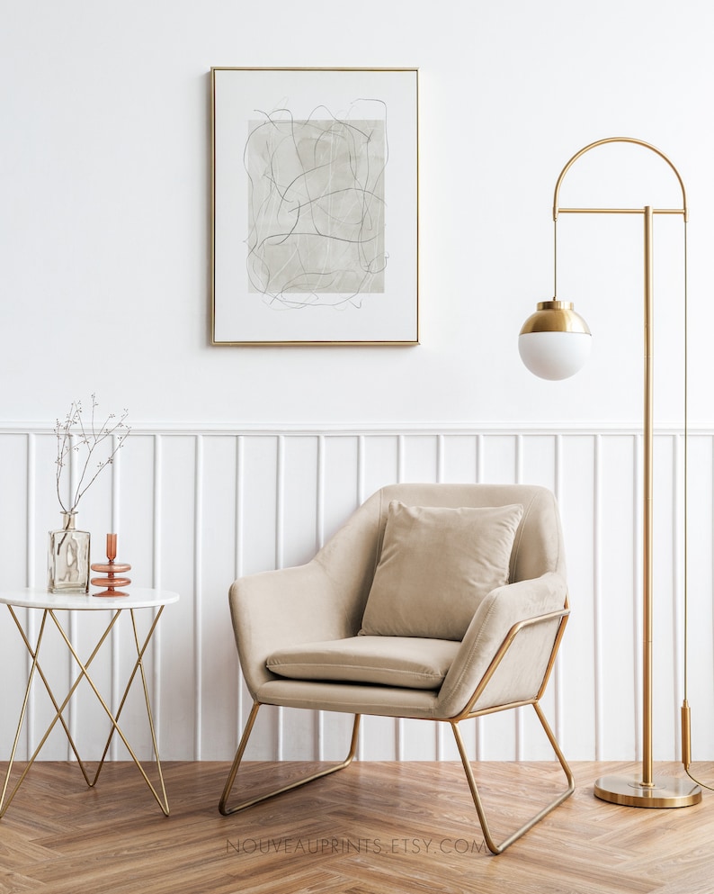 K&ouml;nnte beinhalten: Ein beiger Sessel mit goldenem Metallrahmen und einem beigen Kissen steht in einem Raum mit wei&szlig;en W&auml;nden und einer goldenen Stehlampe mit wei&szlig;em Globus. Ein kleiner wei&szlig;er Tisch mit goldenem Metallrahmen steht im Vordergrund mit einer Vase mit getrockneten Zweigen und einem Kerzenhalter.
