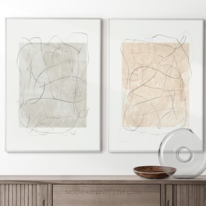 K&ouml;nnte beinhalten: Zwei gerahmte abstrakte Kunstdrucke mit minimalistischem Design. Die Drucke zeigen einen beige-grauen Hintergrund mit schwarzen Linien. Die Drucke h&auml;ngen an einer wei&szlig;en Wand &uuml;ber einem Holzschr&auml;nk.