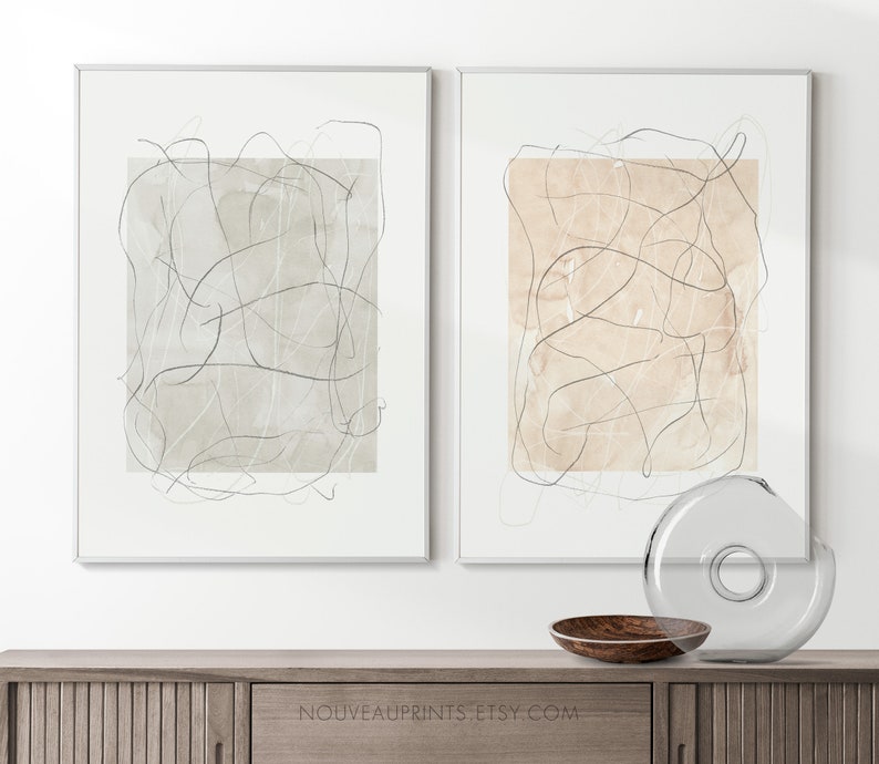 K&ouml;nnte beinhalten: Zwei gerahmte abstrakte Kunstdrucke mit minimalistischem Design. Die Drucke zeigen einen beige-grauen Hintergrund mit schwarzen Linien. Die Drucke h&auml;ngen an einer wei&szlig;en Wand &uuml;ber einem Holzschr&auml;nk.