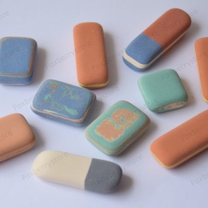 May include: Assortment of ten rectangular rubbers in various colours, including blue, green, orange, and white. Some rubbers have a marbled pattern, while others have a solid colour. The rubbers are arranged on a white surface.