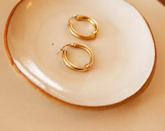 May include: Two gold hoop earrings with a textured design, displayed on a white ceramic plate with a brown rim. The earrings have a polished finish and a classic, elegant style. The earrings are a perfect accessory for any outfit.