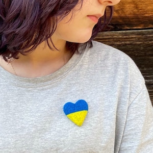 May include: A person wearing a grey shirt with a blue and yellow heart-shaped pin. The pin is in the shape of the Ukrainian flag.