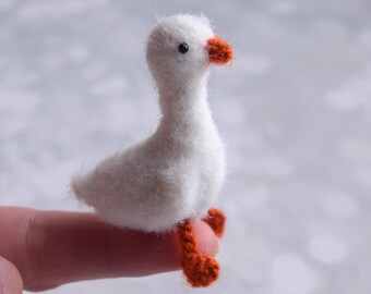 Può includere: Una statuetta di oca bianca in miniatura con becco e zampe arancioni. Il piccolo uccello è fatto di un materiale morbido e sfocato ed è posato su un dito. L'oca è un affascinante oggetto decorativo.