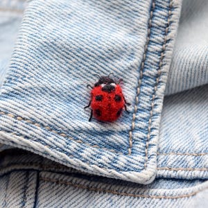 May include: A red and black ladybug pin is attached to a light blue denim jacket. The ladybug has a red body with black spots and a black head. The denim jacket has visible stitching and a classic design.