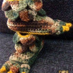 May include: Crocheted slippers with a camouflage pattern and yellow trim. The slippers have a pointed toe and a textured design.