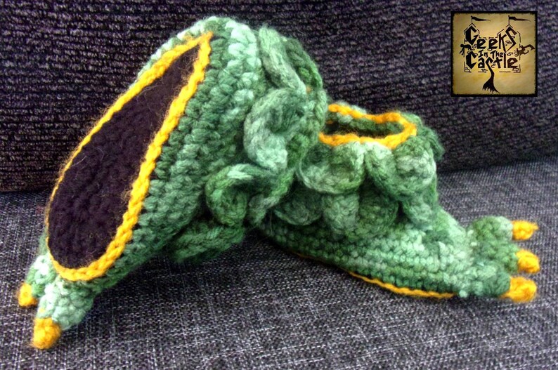 May include: Green and yellow crocheted slippers shaped like a dragon's foot. The slippers have a black felt lining and yellow toes.